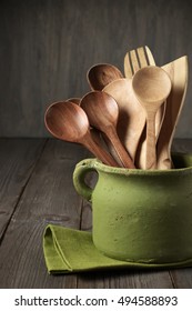 Wooden Cooking Utensils Set In Rustic Handmade Rough Green Ceramic Pot With Napkin On Wooden Table.