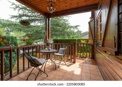 A Wooden Cottage With Beautiful Porch And Table With Two Chairs. Cozy And Nice House With Many Plants And Trees On Sunny Day In European Village.