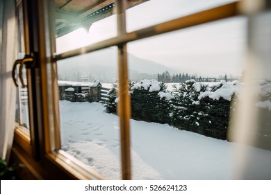 Wooden Cottage House Window. Beautiful Snowy Winter Morning Behind The Chalet Window. Beautiful Mountain View. Sunshine And Sparkling Snow On Fir Trees In The Cabin Backyard