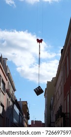 A Wooden Crate Or Box Suspended In Mid Air Hanged By A Chain Attached To Industrial Crane, In Between Two Rows Of Building, On A Sunny Day With Blue Sky.