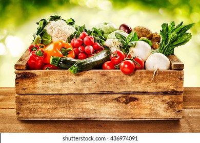 Wooden crate of farm fresh vegetables with cauliflower, tomatoes, zucchini, turnips and colorful sweet bell peppers on a wooden table outdoors in sparkling sunlight on greenery