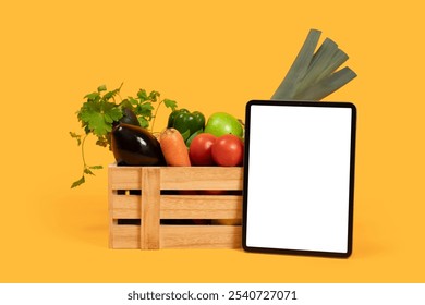 A wooden crate is filled with a variety of fresh vegetables, including tomatoes, eggplant, and leeks, positioned next to a blank tablet against a vibrant yellow backdrop. - Powered by Shutterstock