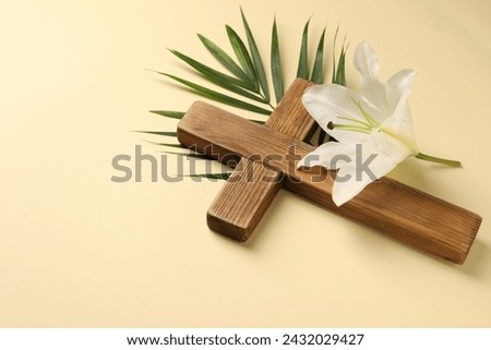 Wooden cross, lily flower and palm leaf on pale yellow background, space for text. Easter attributes