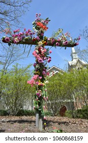 Wooden Cross Outside, Decorated With Flowers And Church In The Background