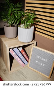 Wooden cube shelf holding stack of used books with modern arts titles, two potted green plants placed on top, framed sign with second hand text leaning against shelf