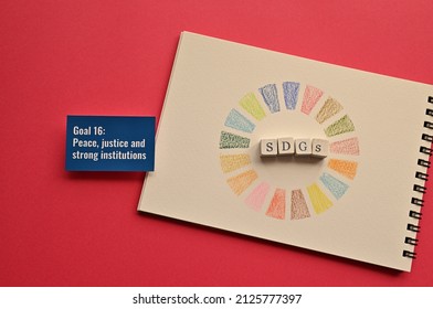 Wooden Cubes Arranged In The Letters Of The SDGs And A Card With The Goal 16:Peace, Justice And Strong Institutions Of One Of The Sustainable Development Goals Written On An Illustration Of Its Symbol