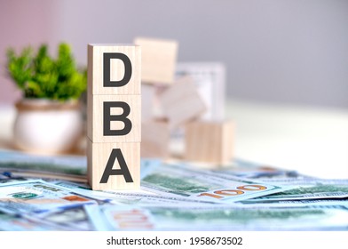Wooden cubes with the letters dba arranged in a vertical pyramid on banknotes, green plant in a flower pot on the background. dba - short for doctor of business administration, business concept.