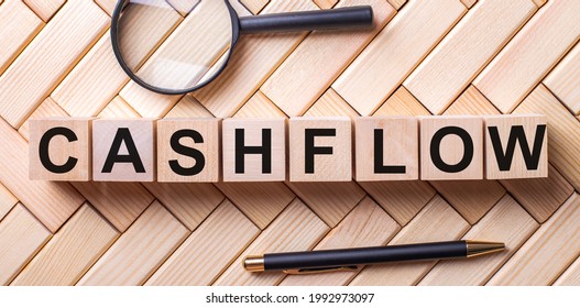 Wooden Cubes With The Word CASHFLOW Stand On A Wooden Background Between A Magnifying Glass And A Pen