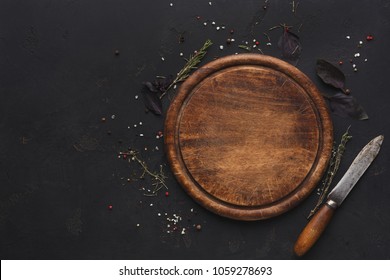 Wooden cutting board with knife and spices on dark background. Rustic catering platter for cutting pizza, bread or meals serving, top view