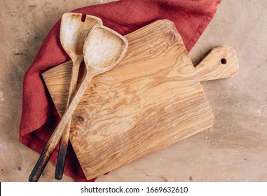 Wooden Cutting Board Over Towel With Utensils On Beige Rustic Kitchen Table. Space For Your Recipe Or Menu. Top View.