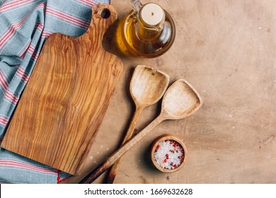 Wooden Cutting Board Over Towel With Utensils On Beige Rustic Kitchen Table. Space For Your Recipe Or Menu. Top View.