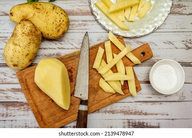 Wooden cutting board with a potato cut into a stick shape to make French fries. Half a peeled potato and a knife on a rustic table. Top view. - Powered by Shutterstock