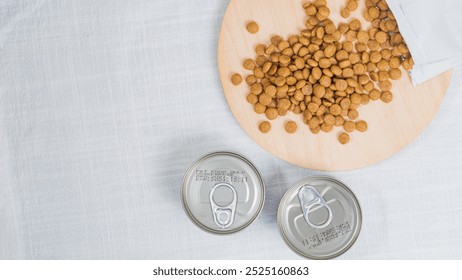 A wooden cutting board with scattered pet food kibble and two open cans of pet food. The background is a light-colored fabric, creating a soft and inviting atmosphere for - Powered by Shutterstock