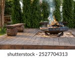 A wooden deck features a circular fire pit with burning logs and smoke. Surrounded by lush greenery and tall evergreens, rustic benches enhance the scene.