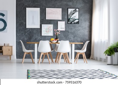 Wooden dining table with white design chairs under window and concrete wall with paintings