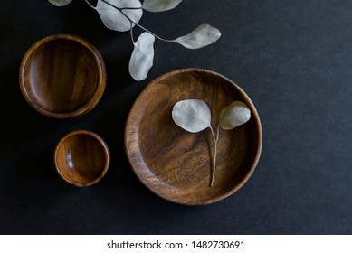 wooden dishes with green cloth napkin and eucalyptus leaves on dark counter, collection of teak dishes at craft fair, beautiful wood bowls and plate on black background, copyspace, copy space