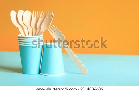 Wooden disposable forks and spoons in a blue paper cup, side view. Eco friendly disposable kitchen utensils on orange background, copy space. Ecological items concept