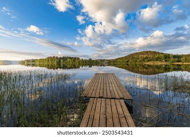 A wooden dock is on a lake with a cloudy sky in the background. The dock is surrounded by tall grass and the water is calm - Powered by Shutterstock