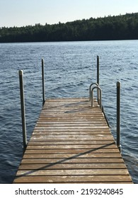 Wooden Dock Out To Lake Water With Shoreline Landscape Of Pine Trees And Blue Sky At Summer Time In Maine 