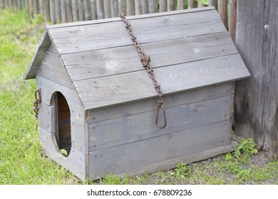 A Wooden Doghouse With An Iron Chain And A Broken Roof, Near A Fence