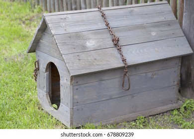 A Wooden Doghouse With An Iron Chain And A Broken Roof, Near A Fence