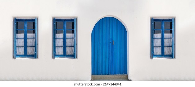 Wooden door closed and three glass windows with open shutters blue color on white wall background. Cyclades island house front view, Greek traditional architecture.