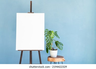 Wooden Easel With Blank Canvas With Monstera Plant On Light Blue Wall Background For Mockup Design. Space For Text