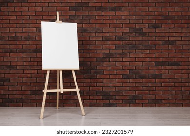 Wooden easel with blank canvas near brick wall indoors. Space for text