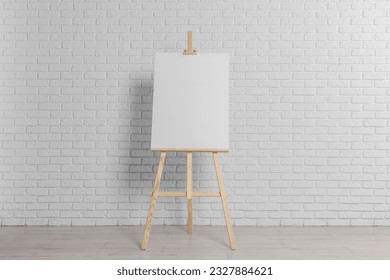 Wooden easel with blank canvas near white brick wall indoors - Powered by Shutterstock