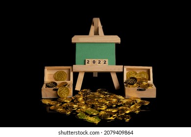 A Wooden Easel With Green Board Written 2022. Left And Right Of Easel Are Wooded Treasure Chest Filled With Gold Coins Spilled On The Floor. A 2022 New Year Financial And Saving Concept. 