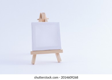 Wooden Easel Miniature With Blank White Square Canvas For Artists And Painters - Mockup. Mini Wooden Stand With Clean Artboard On White Background, Copy Space