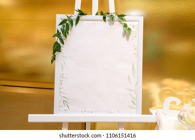 Wooden Easel With White Empty Board And Fresh Green Sprigs Standing Near Gold Wall In Restaurant, Copy Space For Text. Table List Or Seating Chart At Wedding Reception