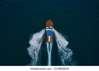 Wooden Expensive Italian Boat With People Fast Movement On The Water Top View. High Speed Open Modern Wooden Boat Moving Fast On Dark Water Aerial View.