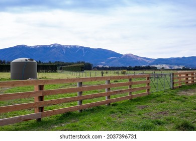 A wooden farm fence with a gate establishes the farm boundary from the country road in Canterbury, New Zealand. A water tank can be seen in the field.