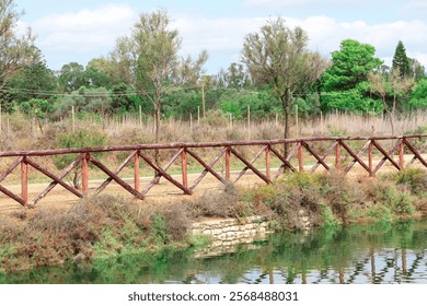 Wooden fence alongside a tranquil pond, surrounded by dry shrubs and trees under a partly cloudy sky. A serene natural setting, blending rustic elements with peaceful greenery - Powered by Shutterstock