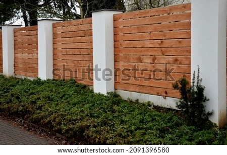 wooden fence made of natural planks. the columns are made of roughly plastered white columns. full fencing made of horizontally placed boards. adjacent to the land is a bed of shrubs