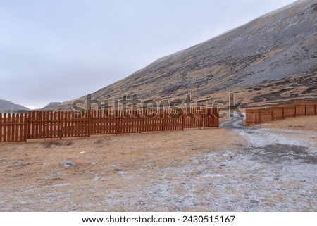 A wooden fence with an open gate crosses a rocky clearing at the foot of a high snow-covered mountain. Valley of the river Naryn-Gol, Altai, Siberia, Russia.