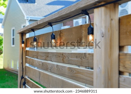 A wooden fence with pine boards and posts. There's a black colored string of outdoor solar powered patio bulbs. The balcony is on the back of a residence. A beige colored shed is in the distance.