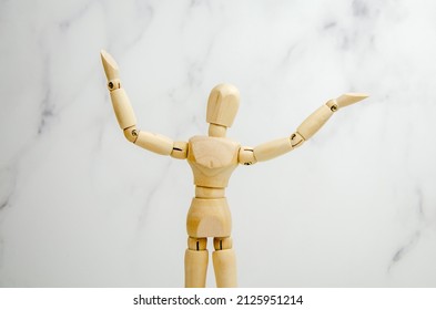 Wooden Figurine Of Man, Raising His Hands Up, Points To An Empty Space. Classic Wooden Mannequin In Demonstration Pose, Standing Against Light Background. Demonstration Of Product, Information, Text