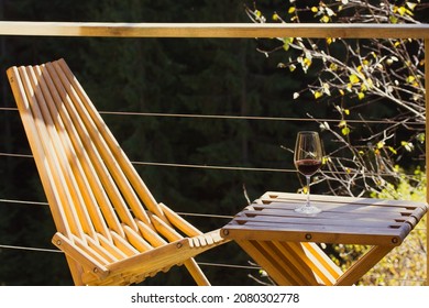 Wooden folding garden furniture - a table and chair - standing on terrace balcony in green woods. A glass of red wine stands on table. Relaxing outdoors at one with nature at autumn. House in forest. 