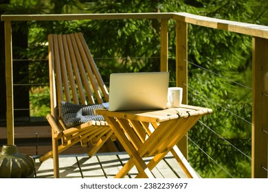Wooden folding table, cup of a tea and laptop for remote work, chairs stand on a balcony of a country house in a green pine forest. Terrace without people. Vacations on a nature. Amazing workplace. 