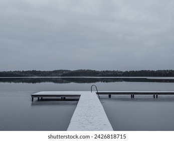 A wooden footbridge covered with snow, swimming place with stairs with first ice, cloudy winter day.