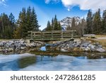 Wooden footbridge in the forest across frozen icy water surface in winter. Snow-covered mountain in background. Johnson lake, Banff National Park, Canadian Rockies, Alberta, Canada.