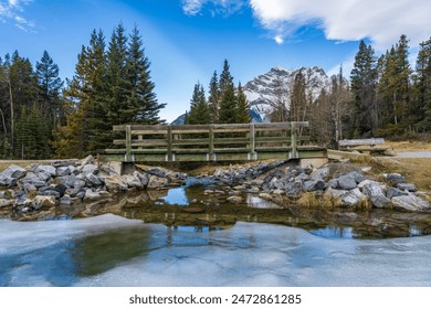 Wooden footbridge in the forest across frozen icy water surface in winter. Snow-covered mountain in background. Johnson lake, Banff National Park, Canadian Rockies, Alberta, Canada. - Powered by Shutterstock