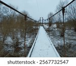 Wooden footbridge over a river on tensioned steel cables with a thin layer of white snow. Long narrow suspension bridge over a river in winter.