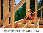 Wooden frame house being built by carpenter. Man wearing glasses hammering nails using hammer and is dressed in a protective helmet and a construction vest. Concept of modern ecological construction.