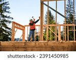 Wooden frame-built home is being erected by carpenter near forest. Bearded man wearing glasses driving in nails with hammer while sporting a safety helmet and a construction vest.