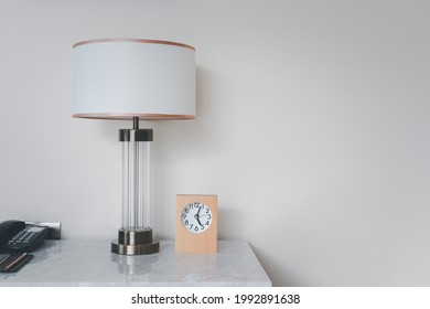  A Wooden Framed Alarm Clock Sits On The Left-hand Side, Resting On A Marble Table And Bedside Lamp.