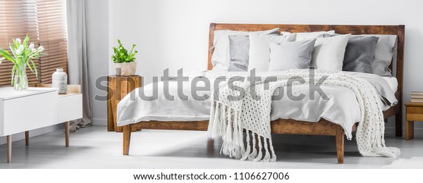 Wooden framed comfortable bed with\
many pillows, blanket and sheets and a sideboard with flowers on\
top in a white stylish bedroom interior. Real photo.\
Panorama.