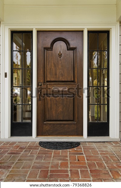 The wooden front door of a home with glass\
panels to each side and a brick porch. The glass is reflecting the\
homes opposite the door. Vertical\
shot.